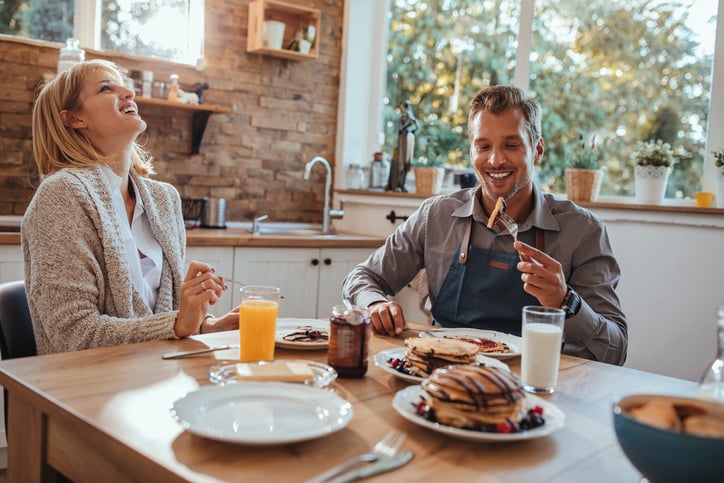 A smiling man and woman eating pancakes at their sunny kitchen table.