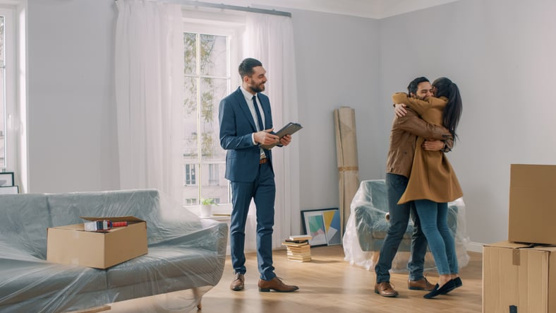 A man and woman hugging in the living room of their new house surrounded by moving boxes standing next to their realtor.