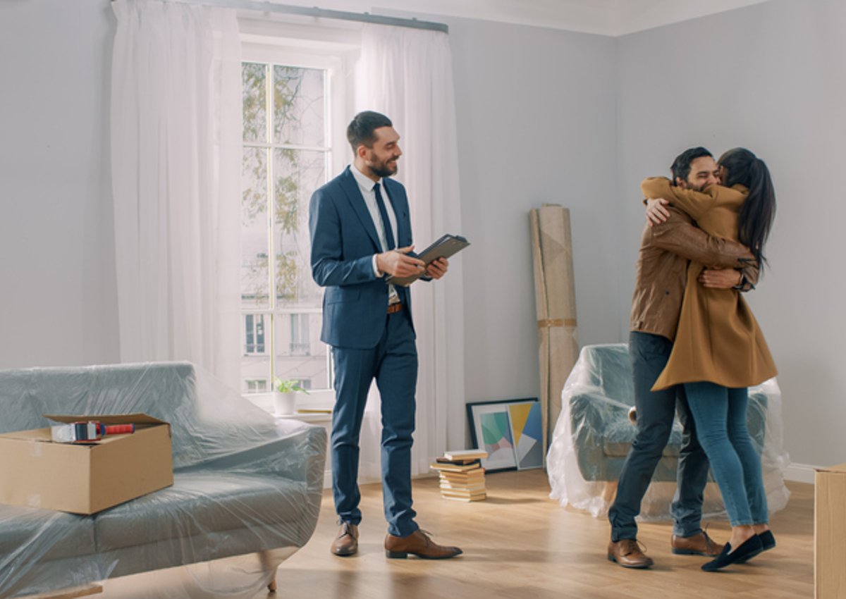 A man and woman hugging in the living room of their new house surrounded by moving boxes standing next to their realtor.