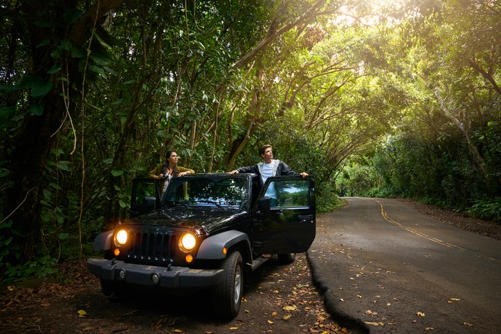 A man and woman standing in the open doors of their car parked on the side of a forest road.