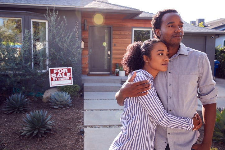 A man and woman standing with their arms around each other in front of their house with a For Sale sign behind them.