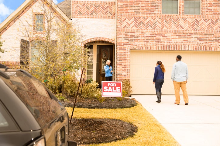 A man and woman meeting their realtor at the front door of a house with a For Sale sign out front.
