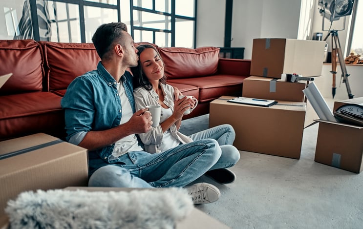 A man and woman smiling while sitting on the floor between moving boxes in the living room of their new home.