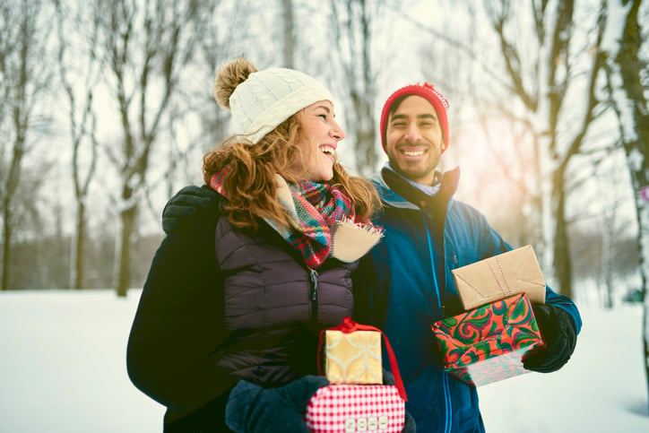 A smiling man and woman walking through a snowy park while holding wrapped gifts.