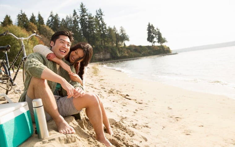 A smiling man and woman sitting next to a bike and ice chest at the beach with their arms around each other.