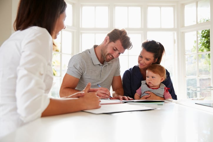 A man sitting next to his wife and baby at a sunny kitchen counter and signing documents from a female mortgage lender.