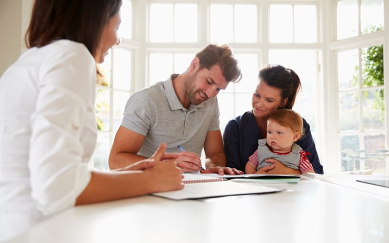 A man sitting with his wife and baby at a sunny kitchen counter and signing documents from a mortgage lender.