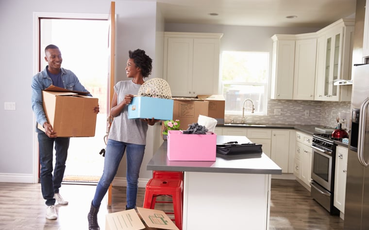 A man and a woman carry boxes into the kitchen of their new home.