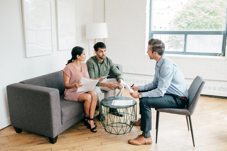 A man and woman sitting on a couch and talking with their realtor while holding paperwork.