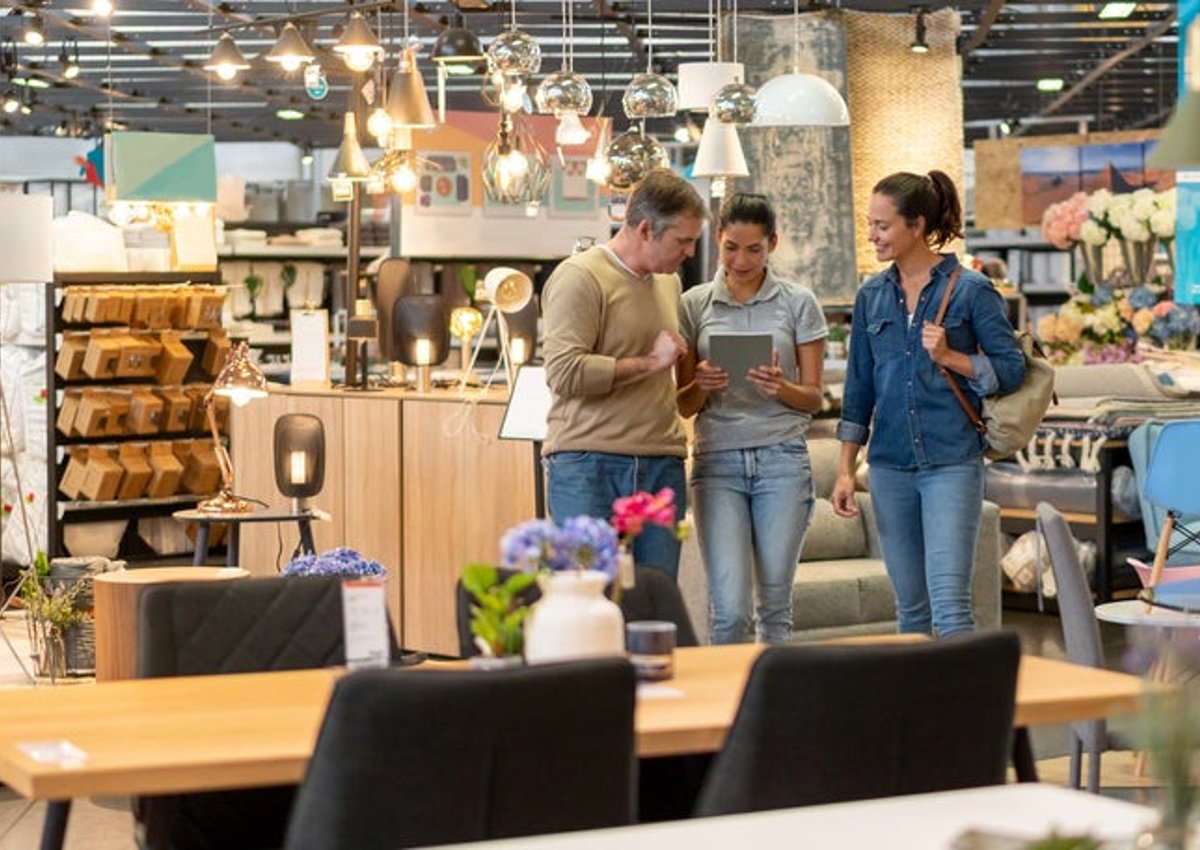 A man and woman walking around a furniture store and looking at a tablet held out by a saleswoman.