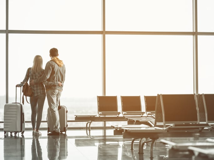 A man and woman standing with their arms around each other and their suitcases next to them at a large airport window.