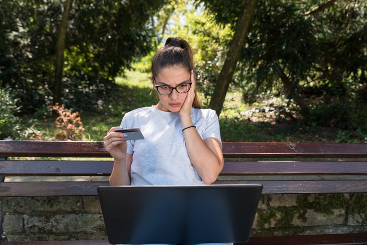 A young woman holding a credit card looks surprised while checking her laptop.