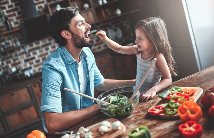 A dad and his young daughter chopping vegetables in the kitchen while she hands him a bite to eat.