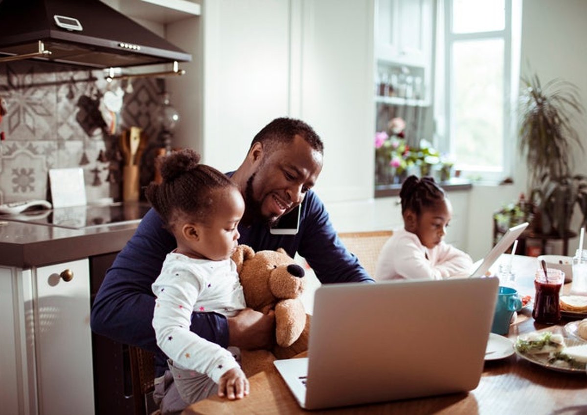 A dad making a phone call and looking at his laptop while sitting at the kitchen table having breakfast with his two young daughters.