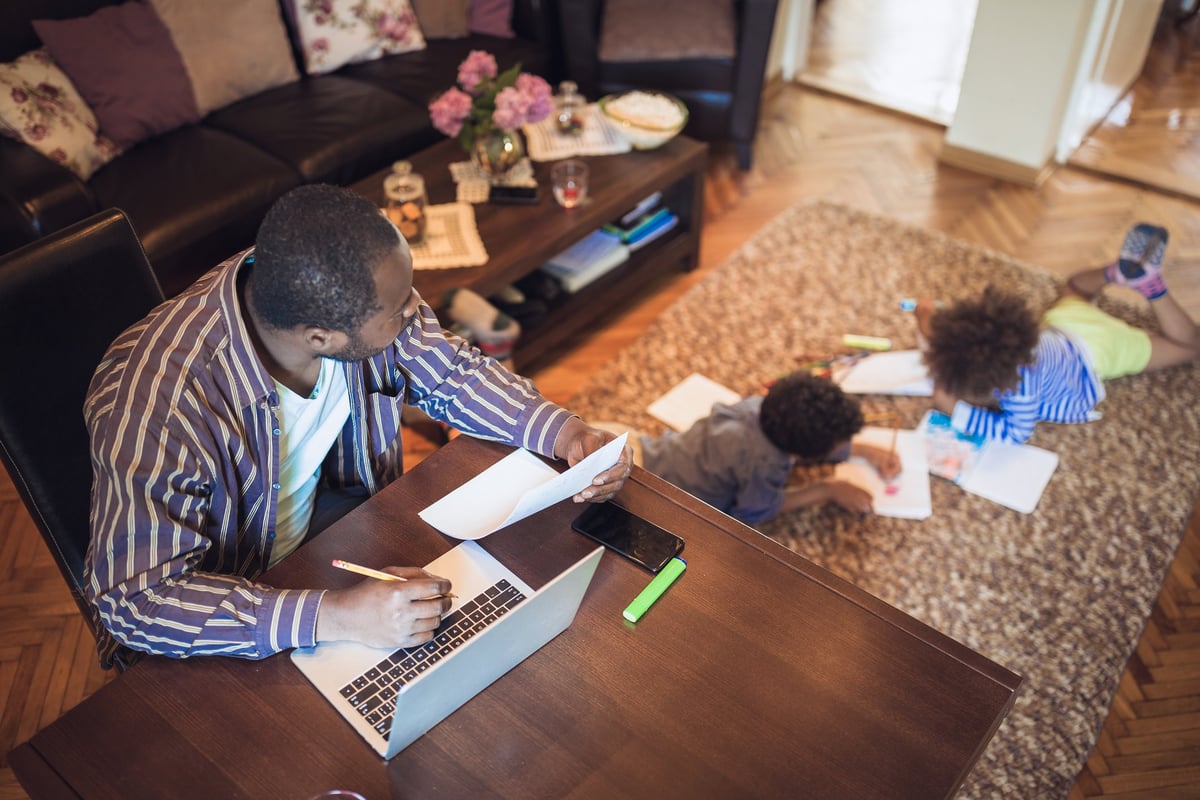 A father working at a table and watching his kids do homework on the floor of the living room.