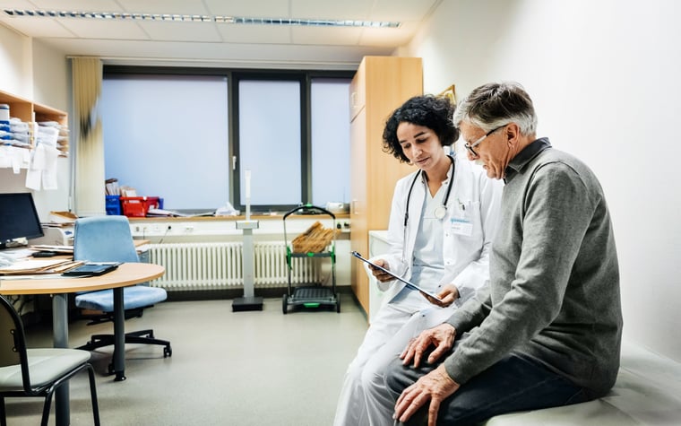 A doctor sitting next to a patient in an exam room and going over test results on a clipboard.