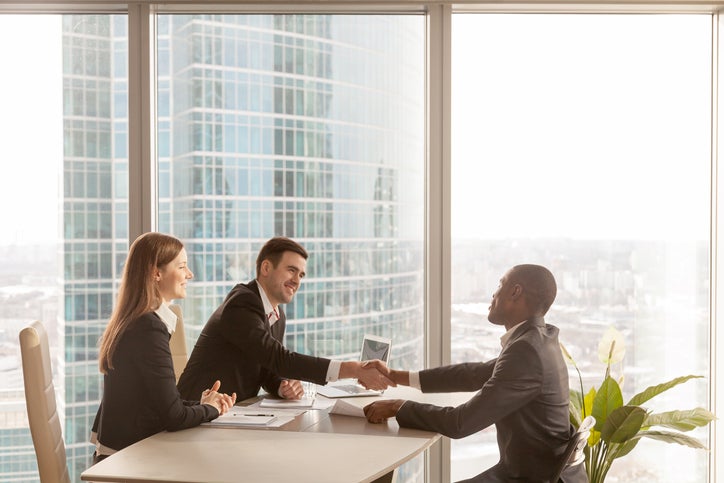 An employee shaking hands with his two bosses across a desk in a sunny high-rise office.