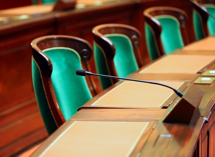 A row of empty seats in front of a long curved desk and microphones in a congress hall.