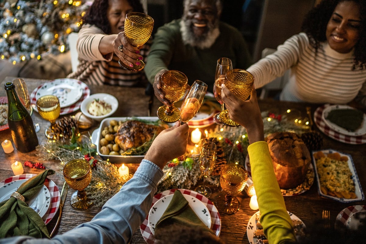 A smiling family toasting their drinks while sitting around a holiday dinner table filled with food.
