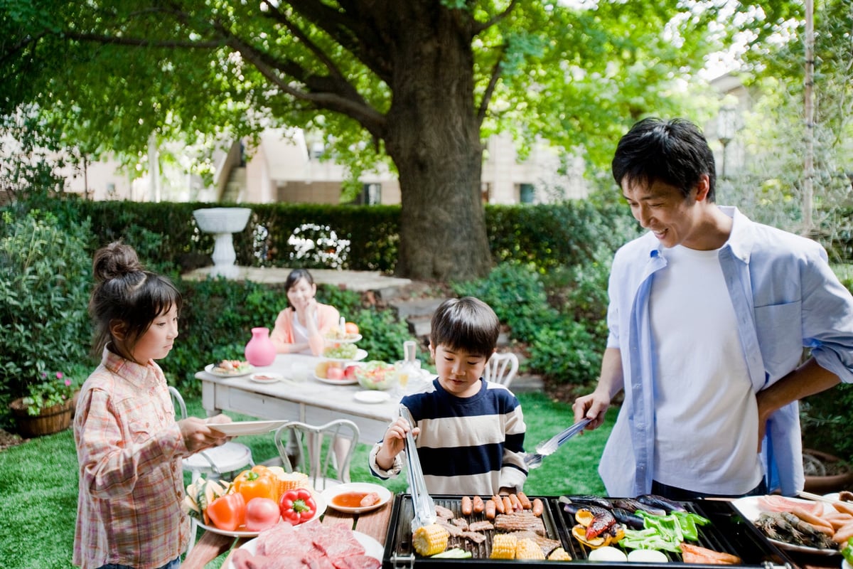 Two parents and two children enjoying a barbecue in their backyard surrounded by green trees.