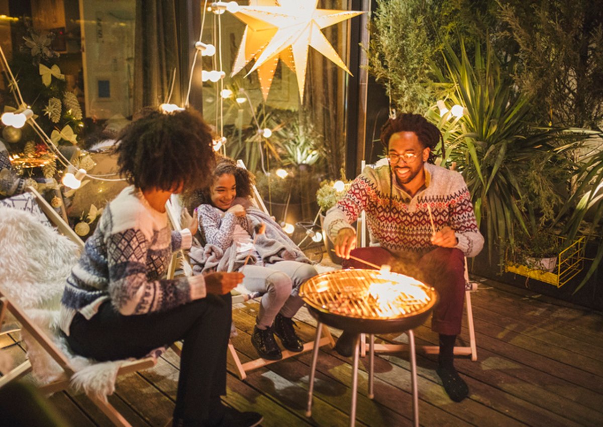 Two parents and their child making smores on their backyard patio decorated with twinkle lights.