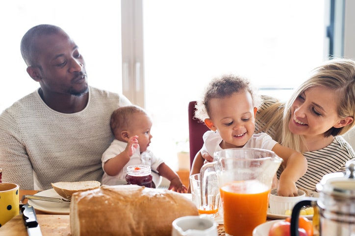 A mom and dad smiling and each holding their toddler and baby while eating breakfast at a sunny table.