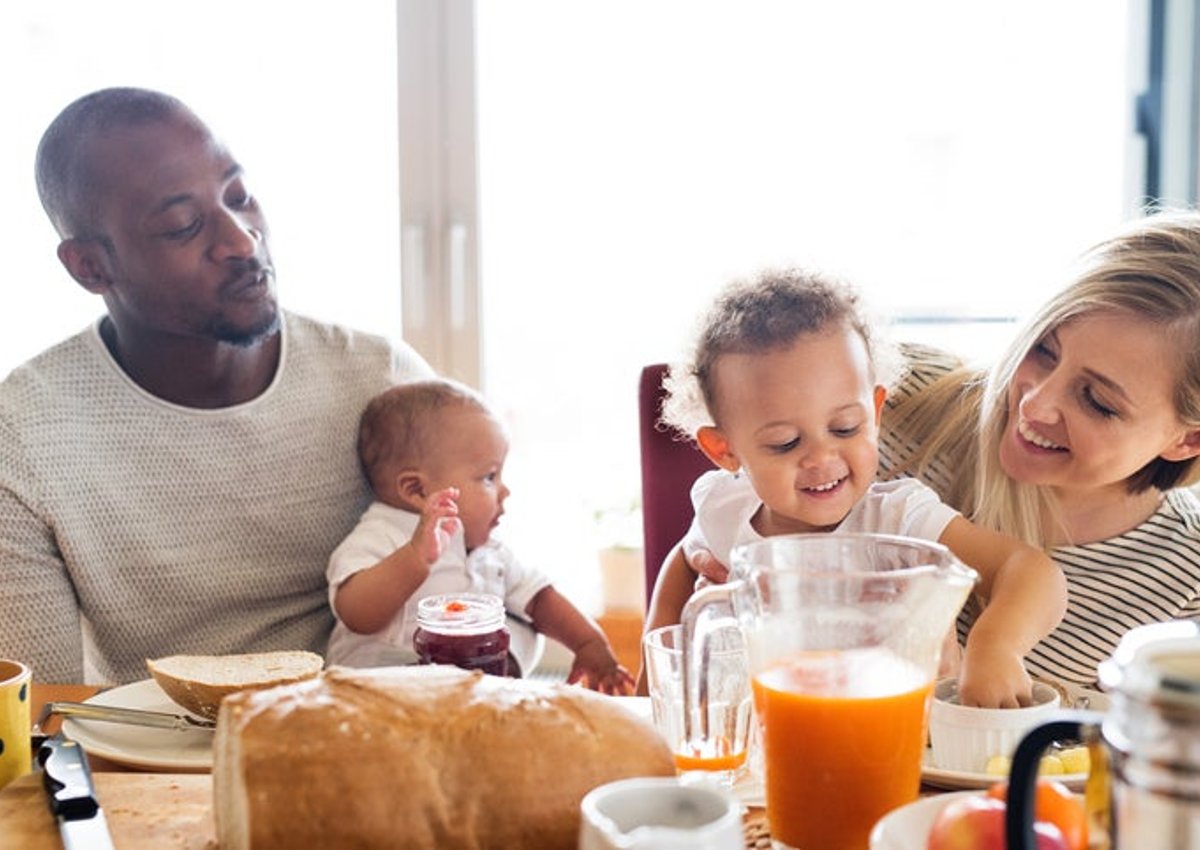 A mom and dad smiling and each holding their toddler and baby while eating breakfast at a sunny table.
