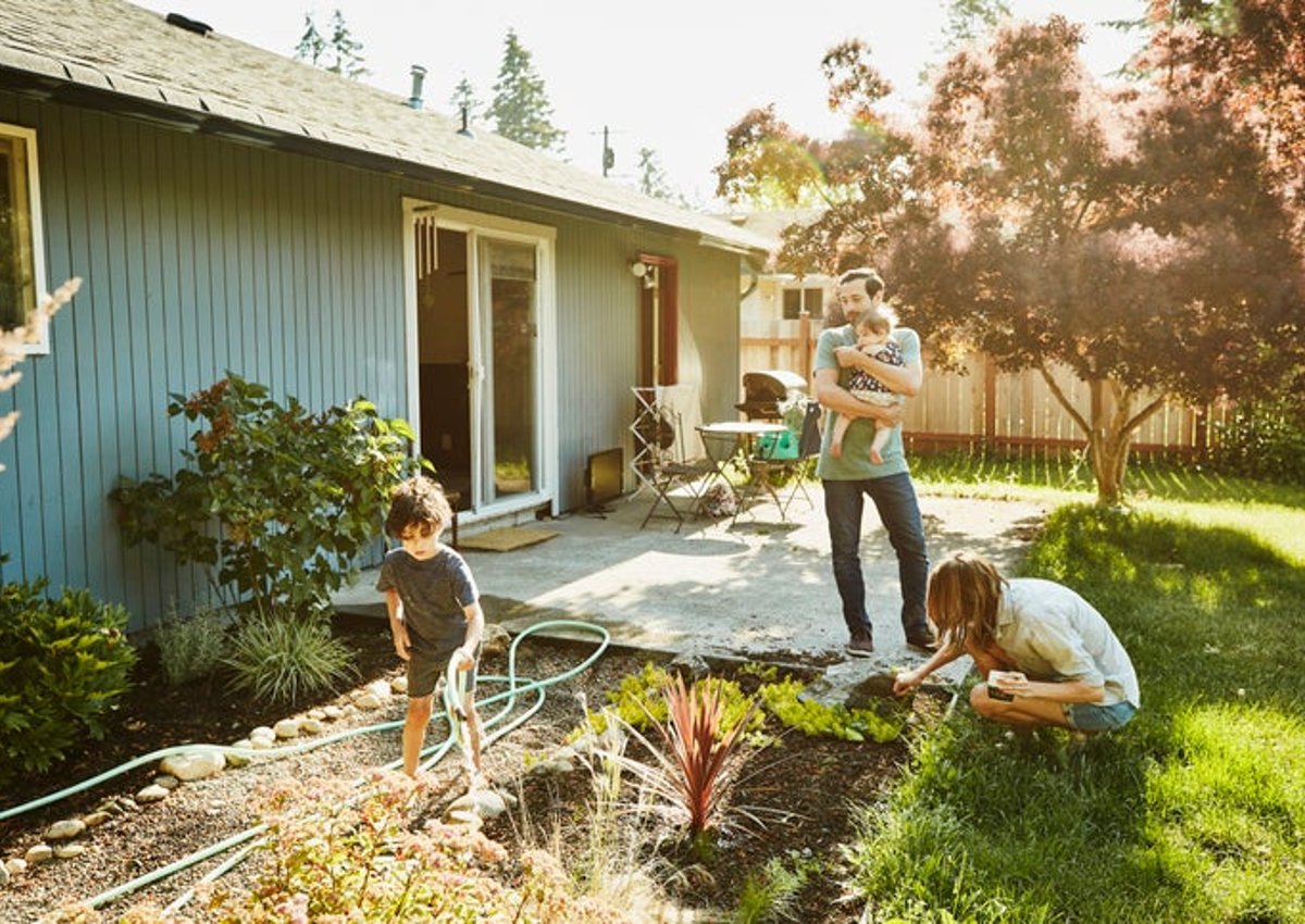 A mom working in the backyard garden with a toddler while the dad walks out of the house holding a baby.