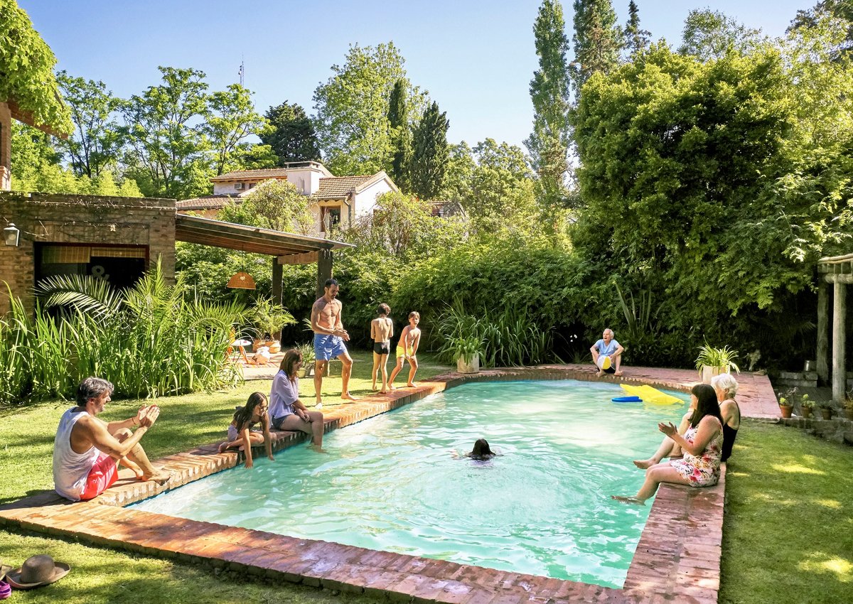 A family playing in a backyard pool surrounded by green bushes and trees.