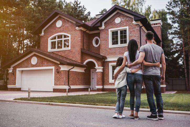 A mom, dad, and their daughter standing outside with their arms around each other and looking at their new home.