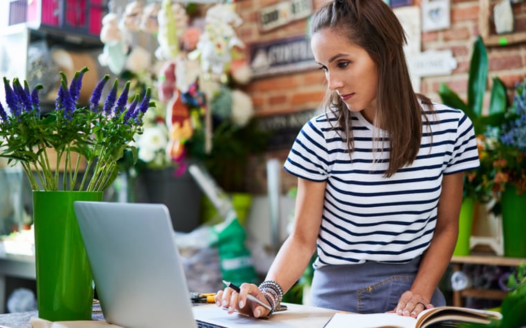 An owner of a flower shop working on a laptop at the counter while writing in a notebook.