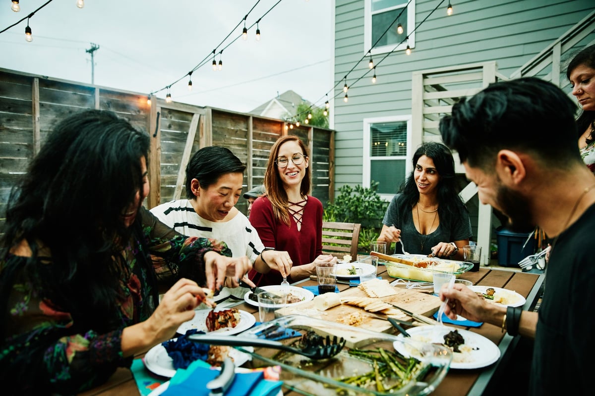 A group of friends eating a meal around a backyard patio table.