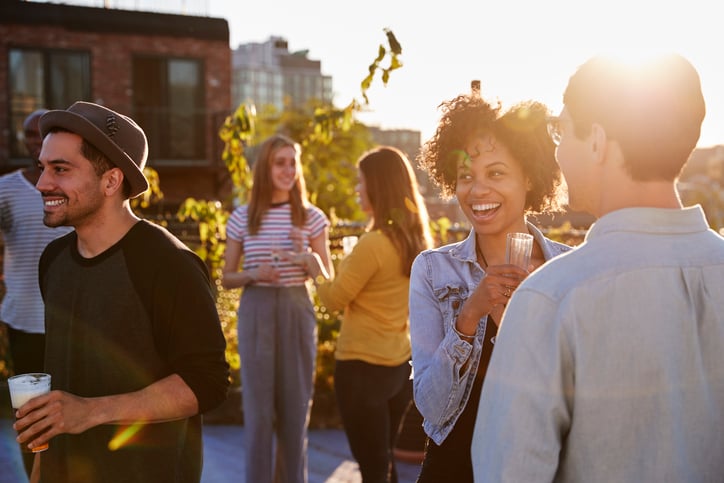 A group of smiling young adults drinking cocktails at a rooftop party at sunset.