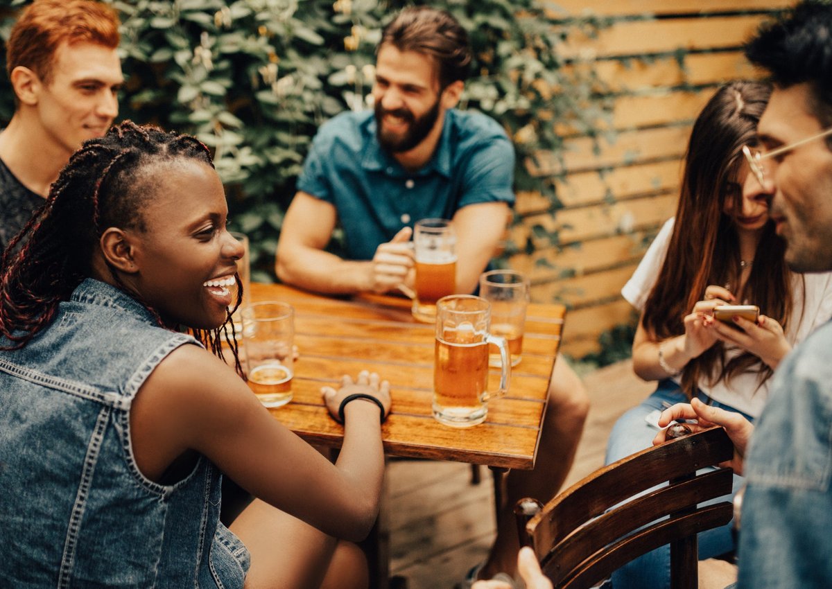A group of laughing friends drinking beers at a restaurant patio table.