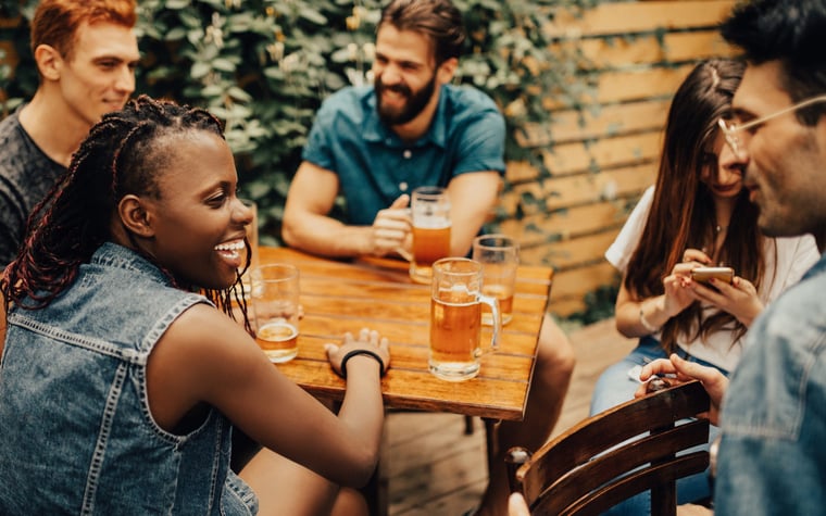 A group of laughing friends drinking beer at a table in a restaurant terrace.