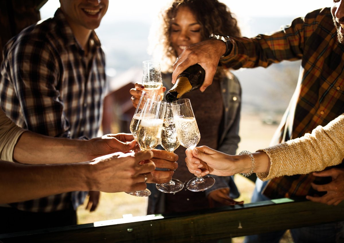 A group of friends hold glasses of champagne to cheer as someone pours the bottle.