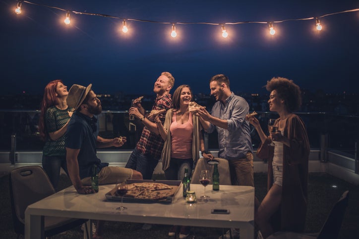 A group of laughing friends sharing pizza and beers on a rooftop at night under decorative string lights.
