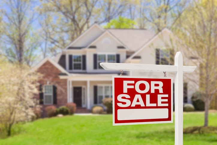 A For Sale sign in front of a two-story house surrounded by trees and a lawn.