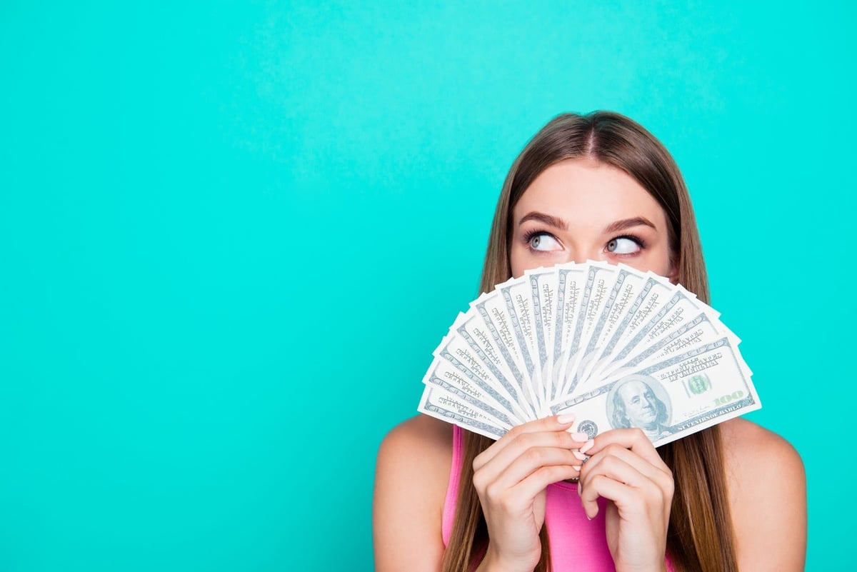 Young woman holding a fan of hundred-dollar bills in front of her face.