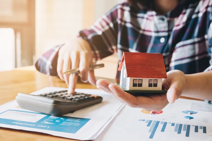 A man with a small house in his hand works a calculator.