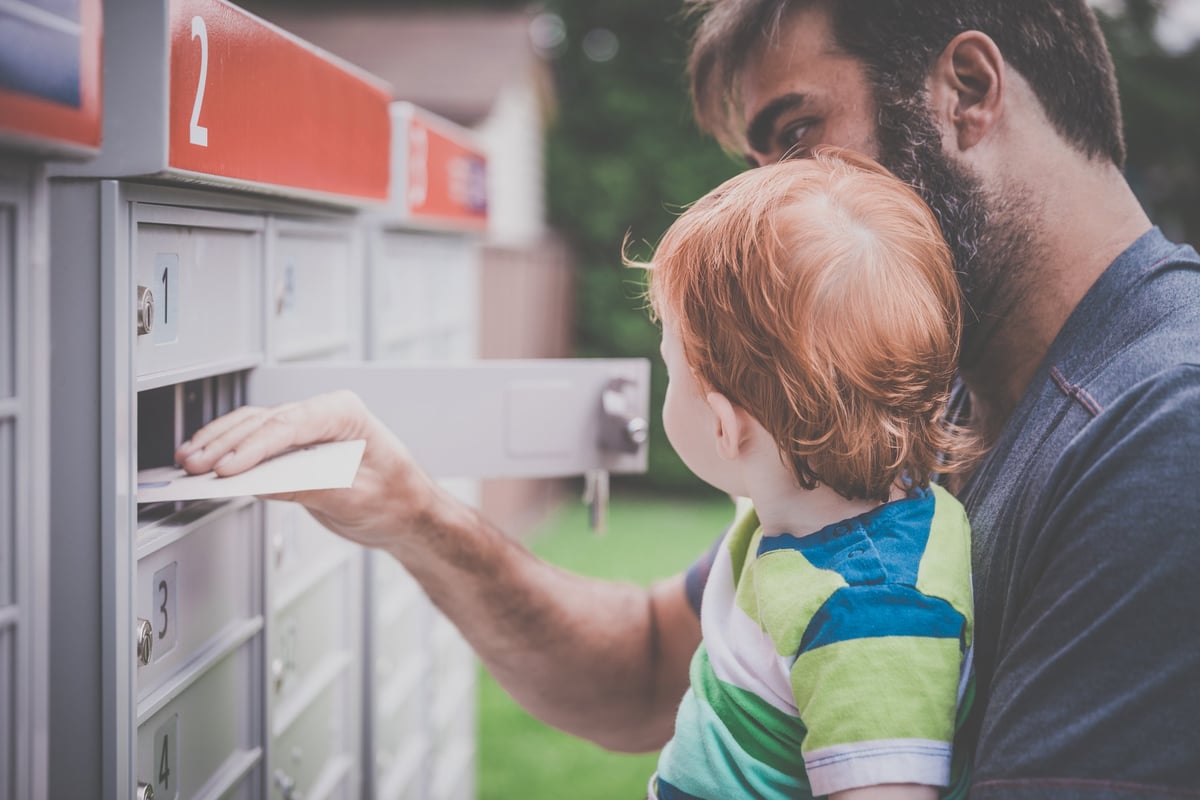 A man holding his baby while opening a mailbox and removing an envelope.