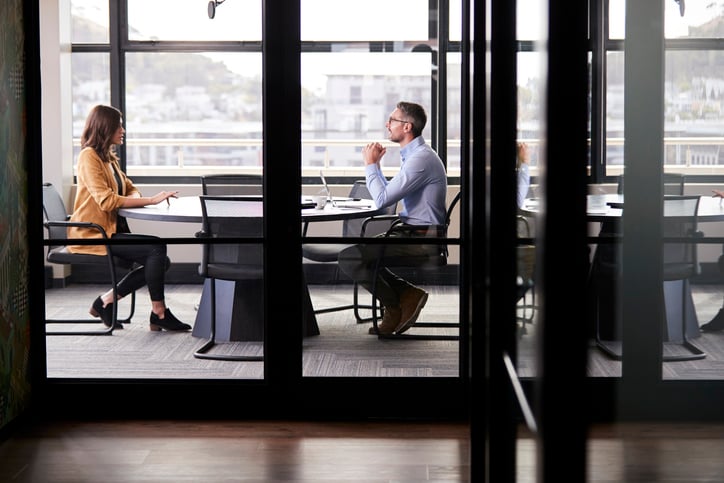 A man and woman sit at opposite ends of a table in a meeting room for a job interview.