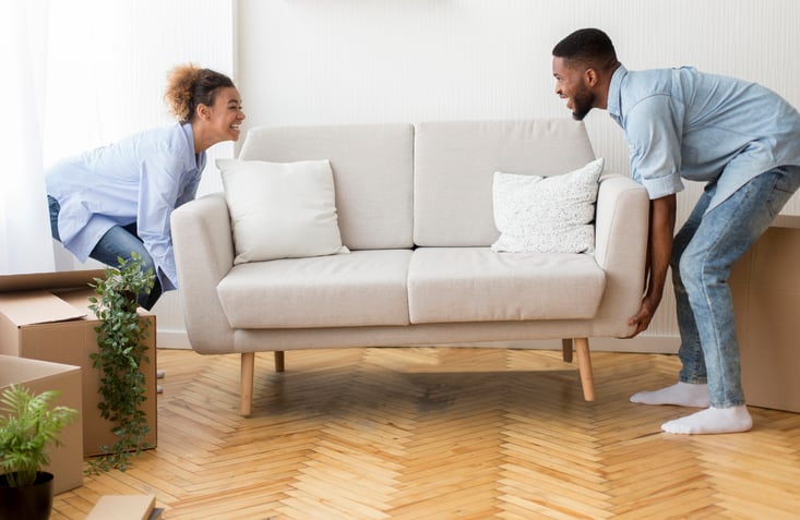 A man and woman moving a new couch into their living room.