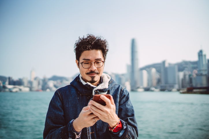 A man looking at his phone while standing along a waterfront with a city skyline behind him.