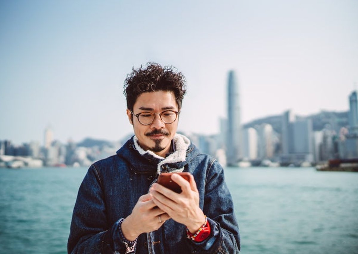A man looking at his phone while standing along a waterfront with a city skyline behind him.