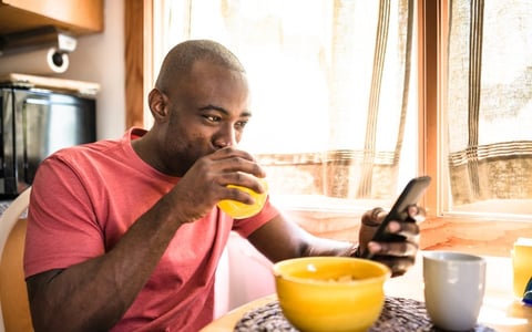 A smiling man drinking juice and looking at his phone while sitting at the breakfast table in front of a bowl of cereal.