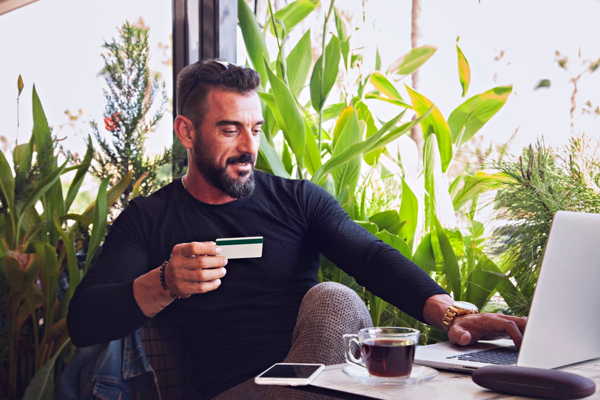 A man sitting in a coffee shop in front of lots of plants working on his laptop and holding a credit card.