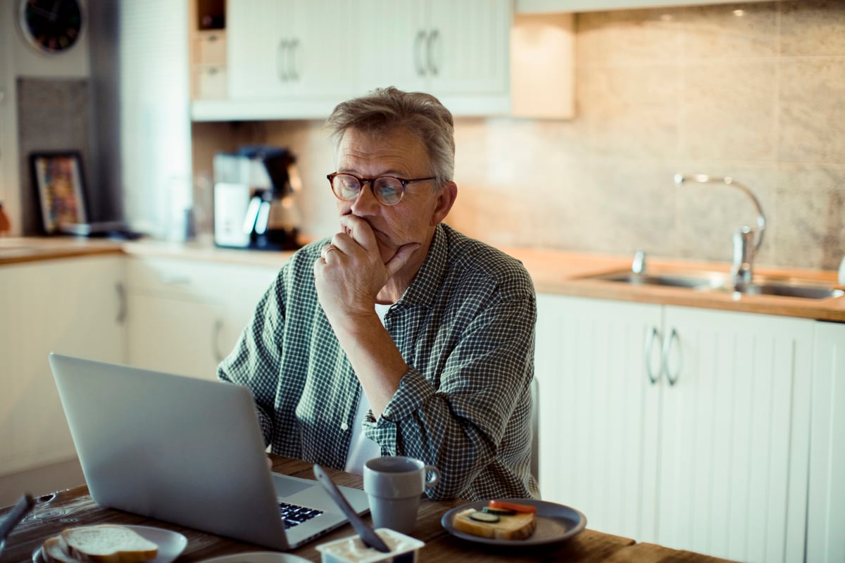 A man holding his chin with his hand while sitting in his kitchen searching on his laptop.