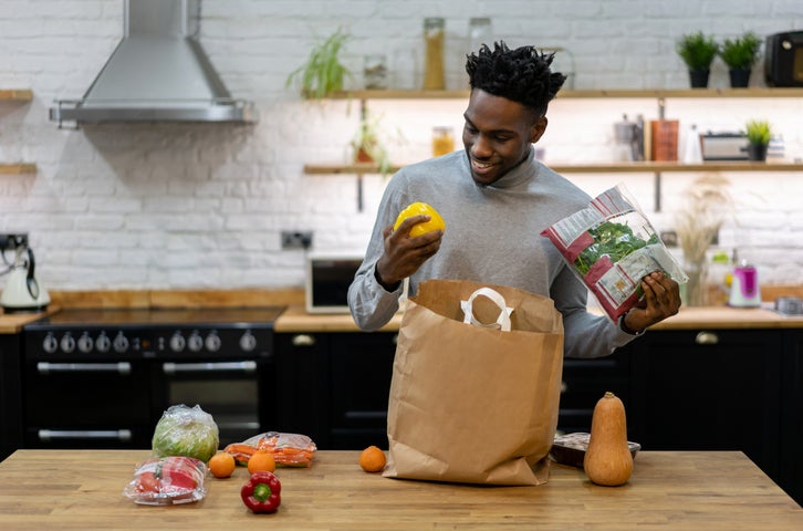 A smiling man standing in his kitchen and unpacking bags of groceries.