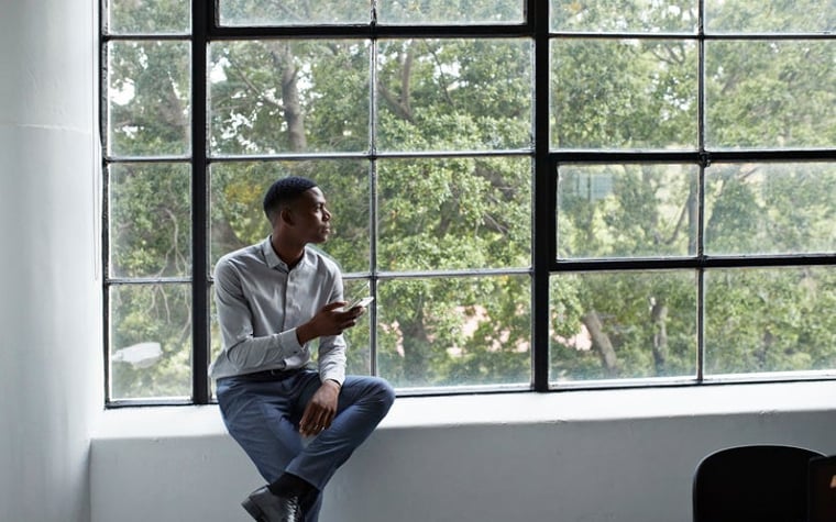Un homme assis à côté d'une grande fenêtre et tenant un téléphone tout en regardant pensivement dehors.